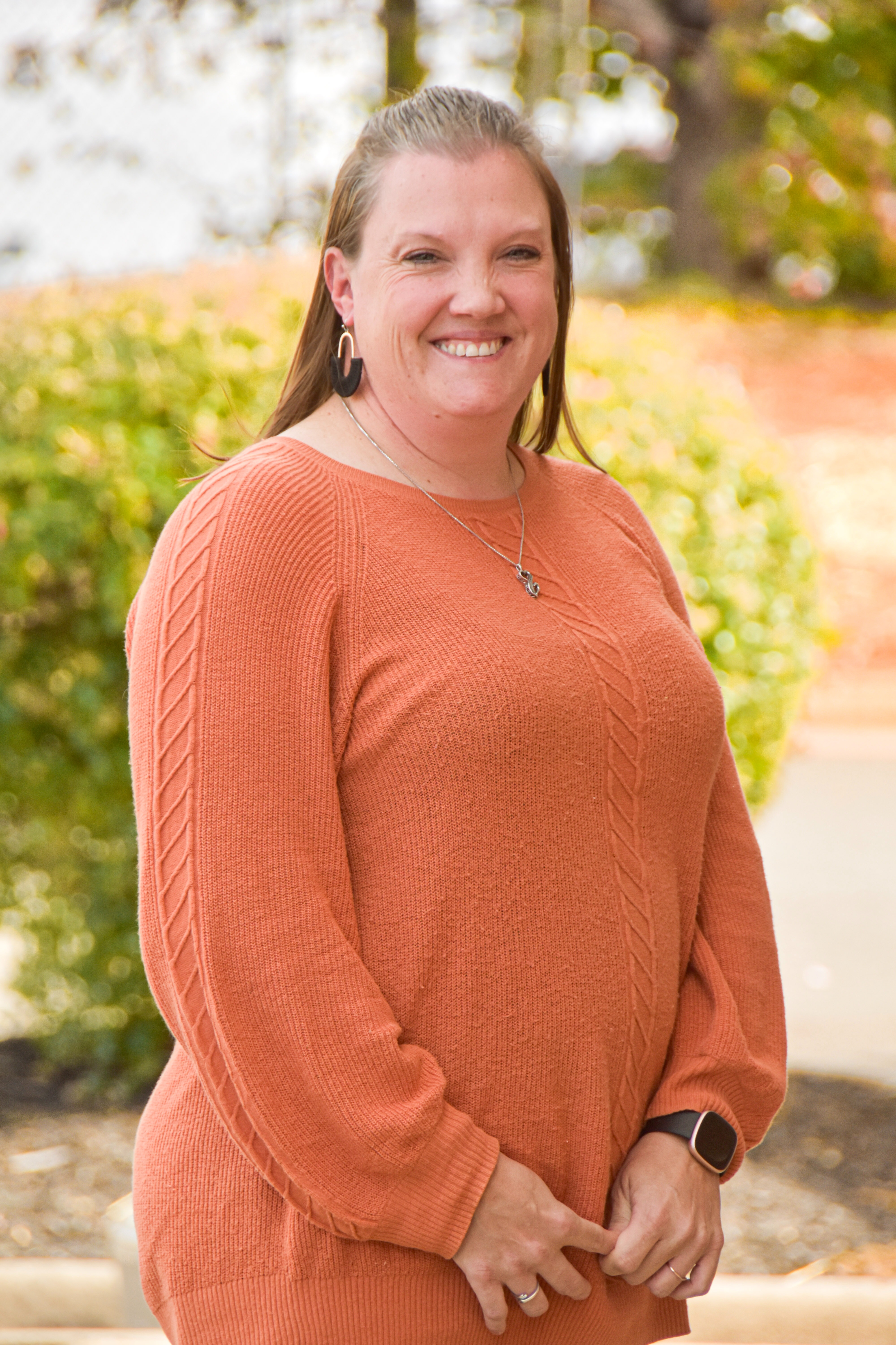 Woman standing outside smiling. She wears an orange sweater and holds her hands in front of her.