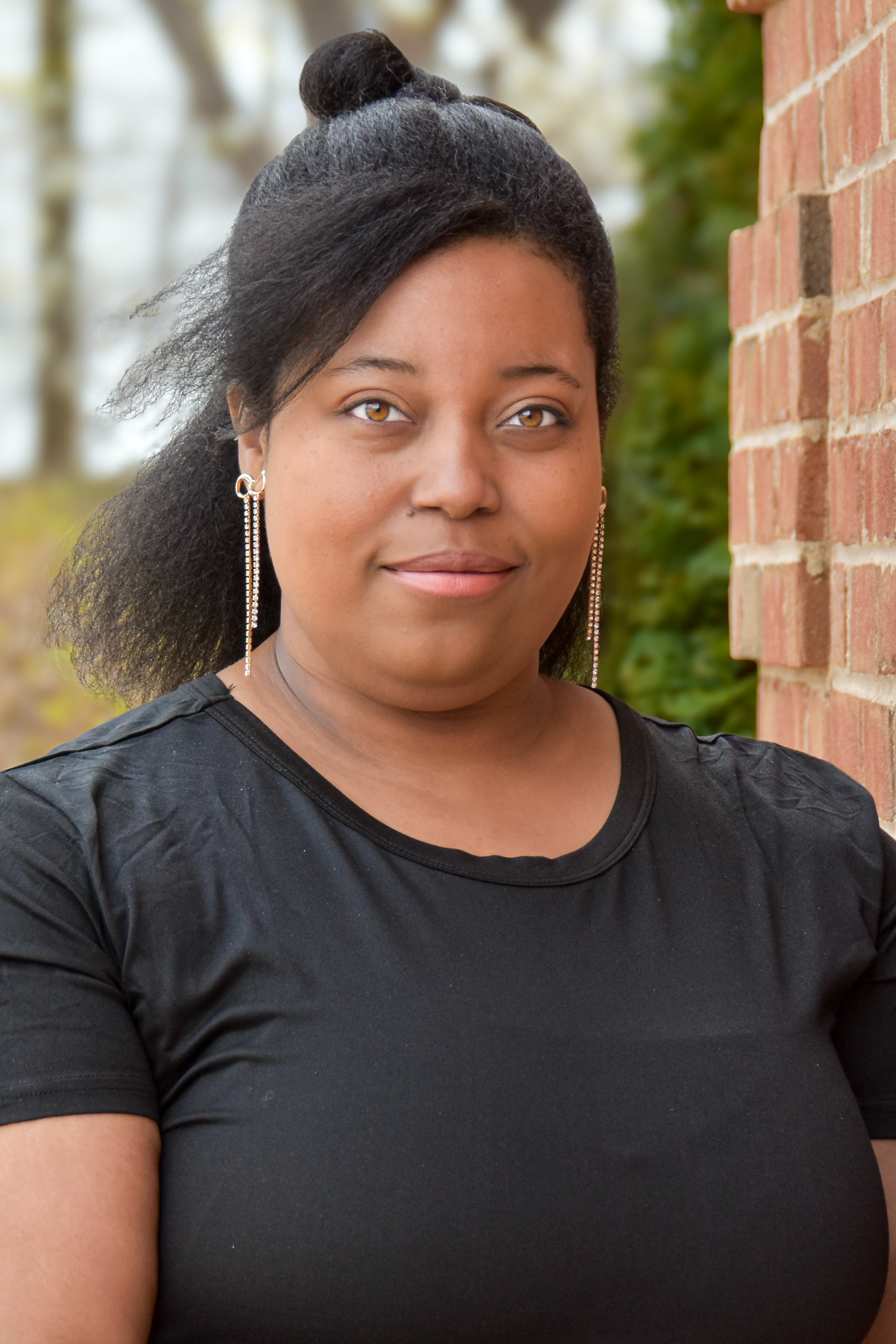 Person with dark hair, wearing black shirt, standing against brick wall, smiling at camera.