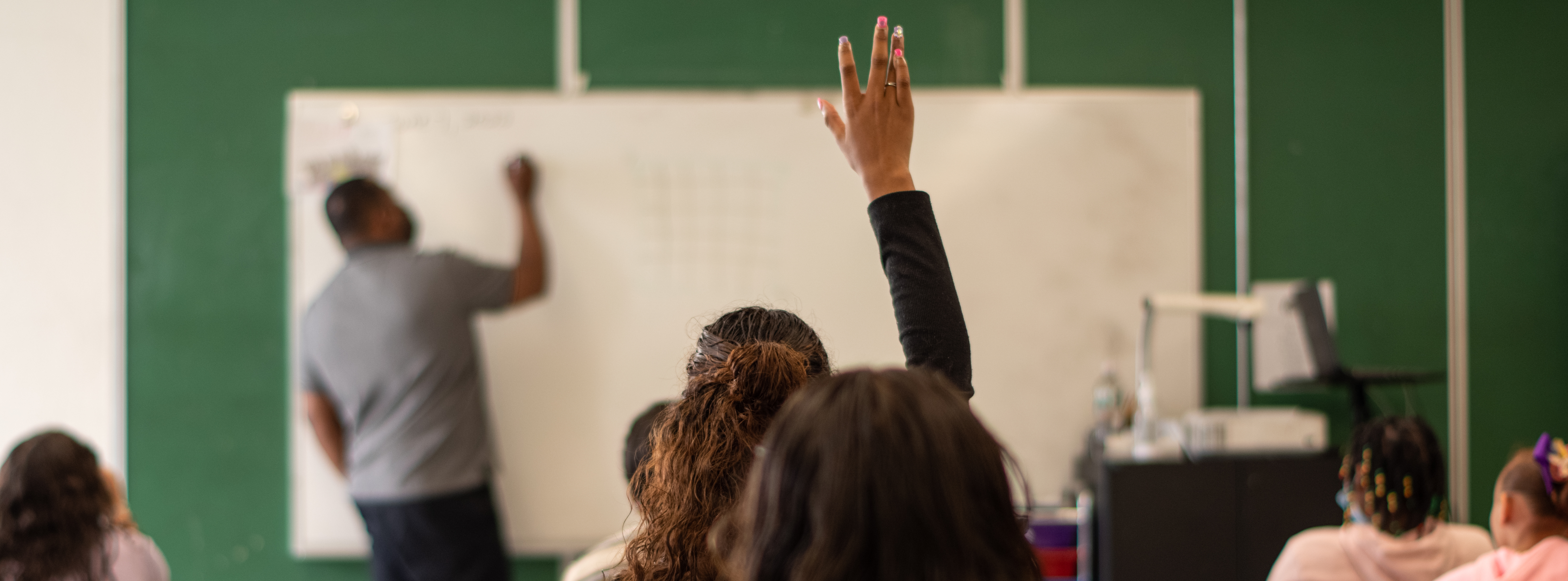student raising her hand in a class while teacher teaching lesson