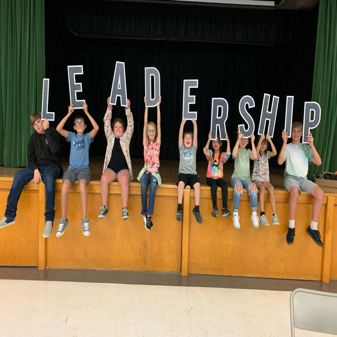 nine students sitting on a stage holding letters that spell out the word leadership