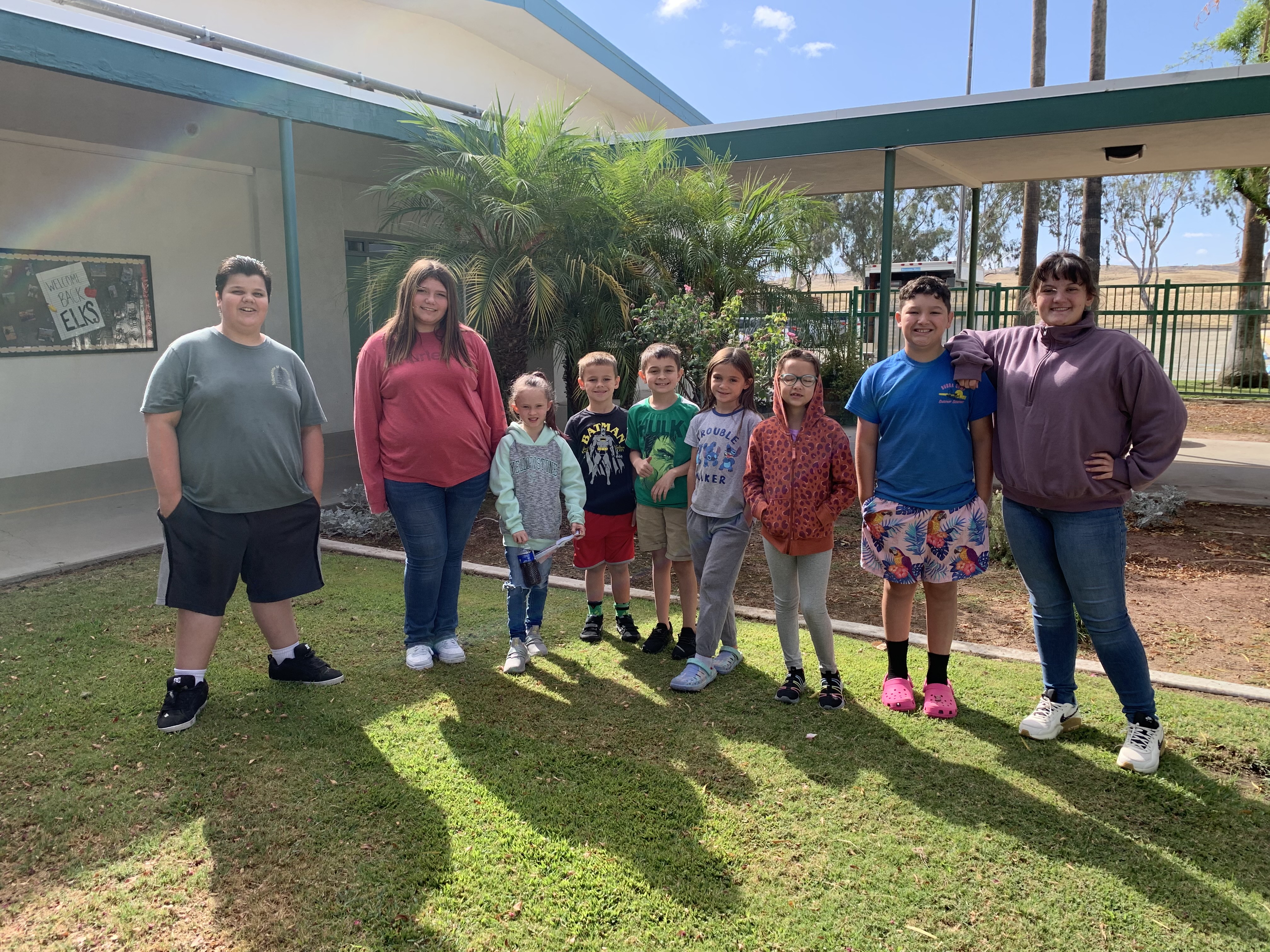 August's Elk of the Month recipients standing on the grass in front of the palm tree. Pictured left to right: Clifford, Mimi, Audrey C., Sirius, Evan M., Bella, Nymeria, Dean, Abbie H. 