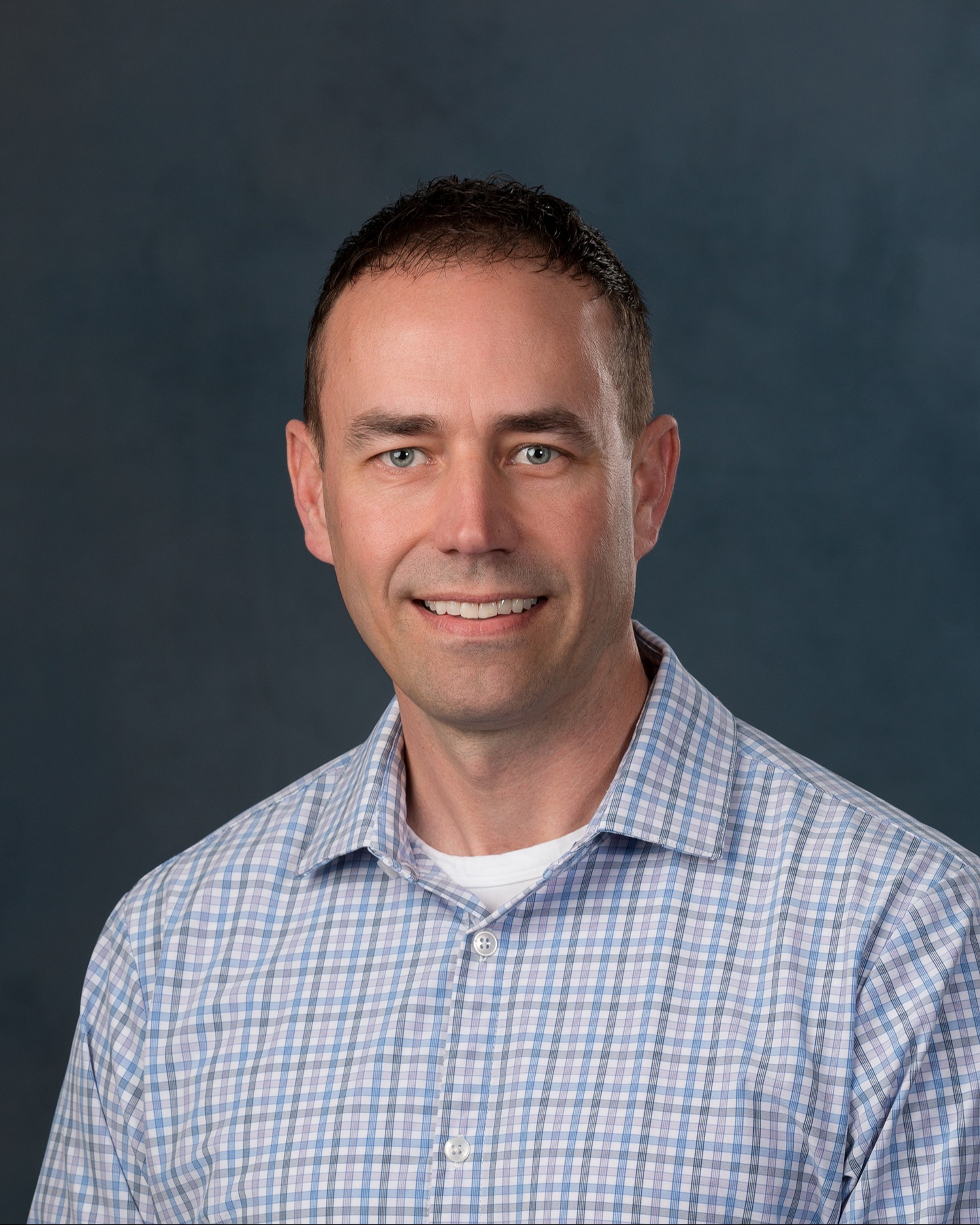 Ryan Brune headshot: man with dark hair wearing a blue plaid button up shirt