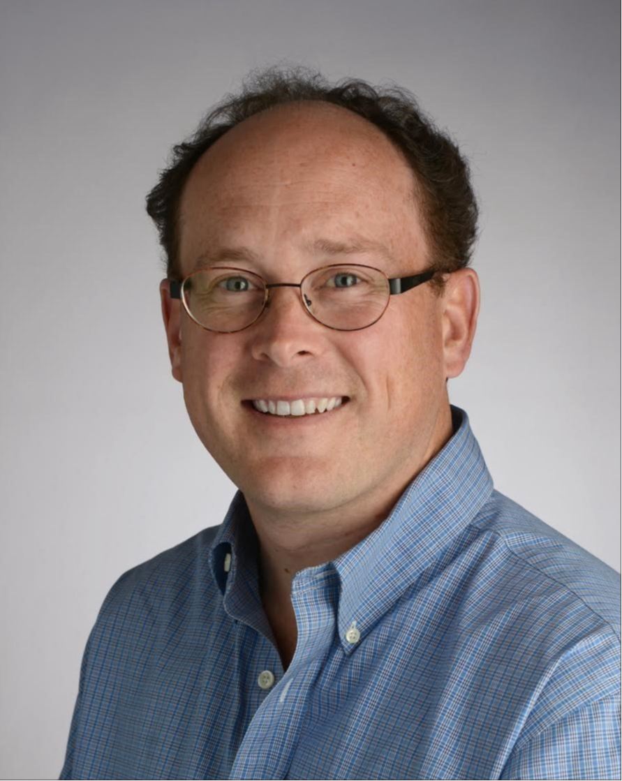 Evan Dean headshot, wearing glasses and a blue and white checked button up shirt