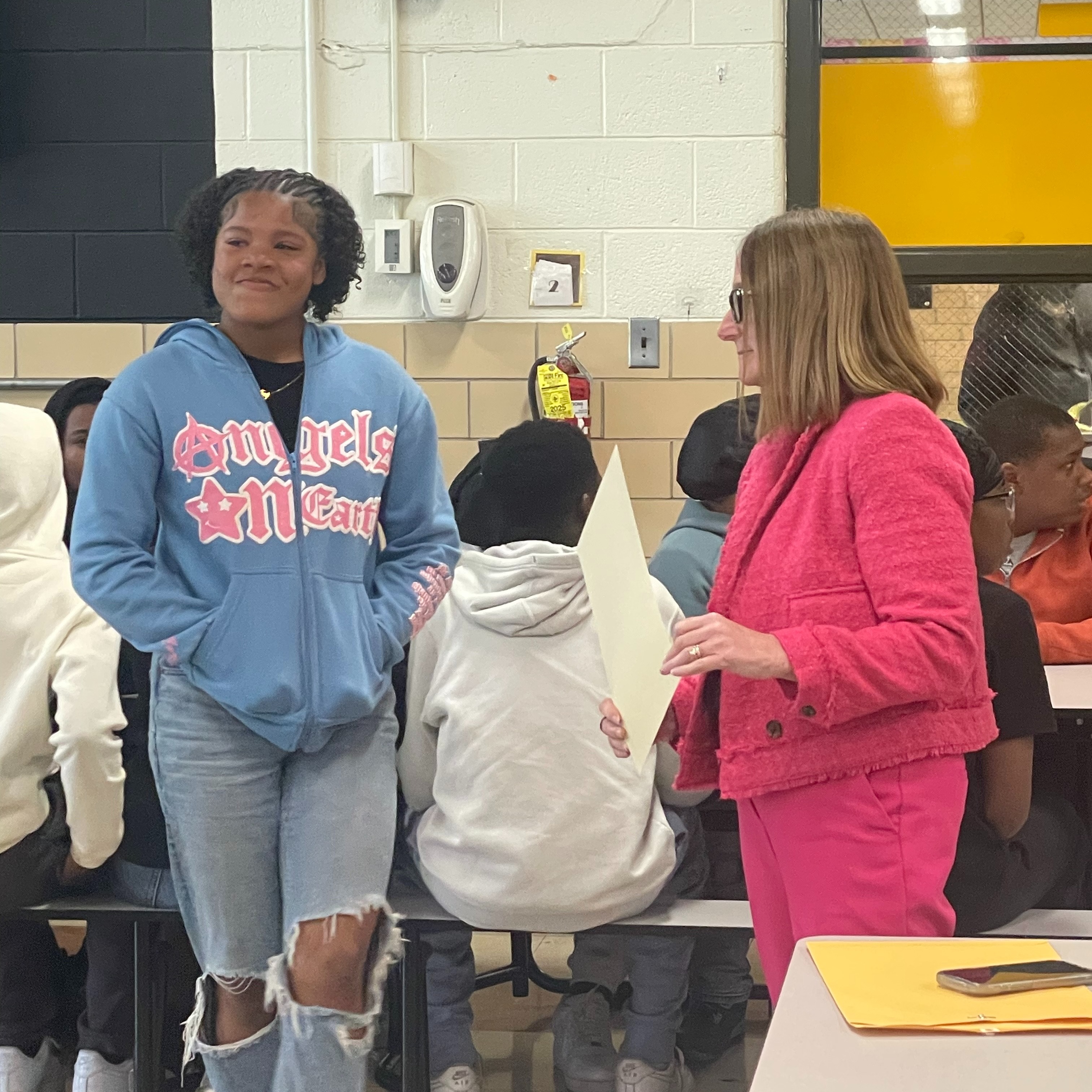 Several students sit at tables. Two adults stand facing each other. One holds a paper. The other wears a blue hoodie.