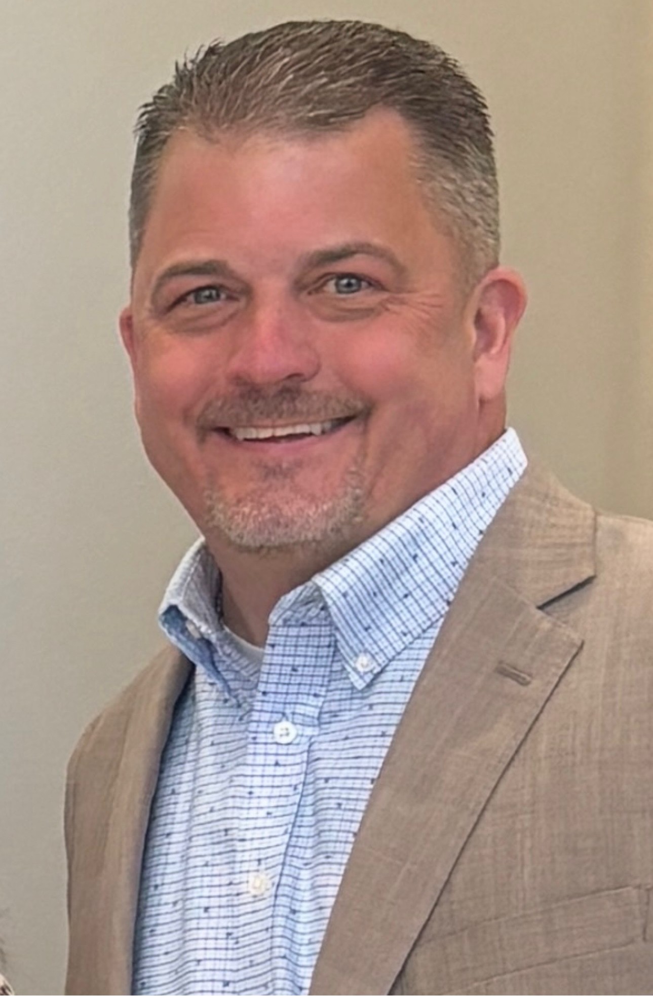 A man in a blue shirt with a tan jacket, short hair, and a beard, smiling for a photo.