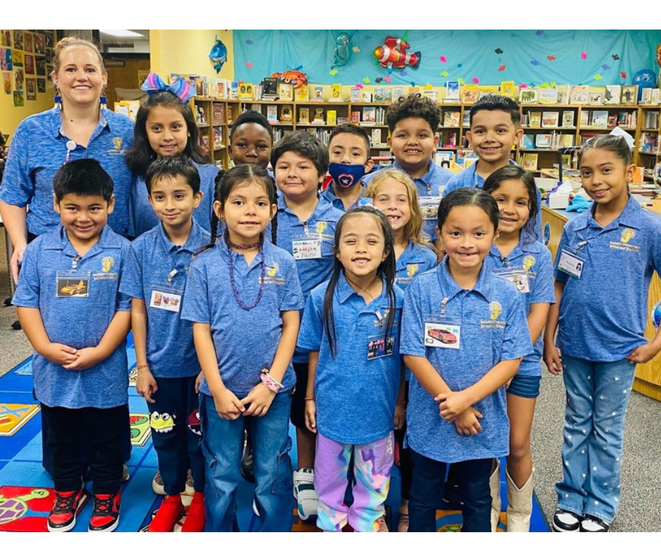A classroom with a teacher and young children in matching blue shirts, standing in front of a bookshelf.