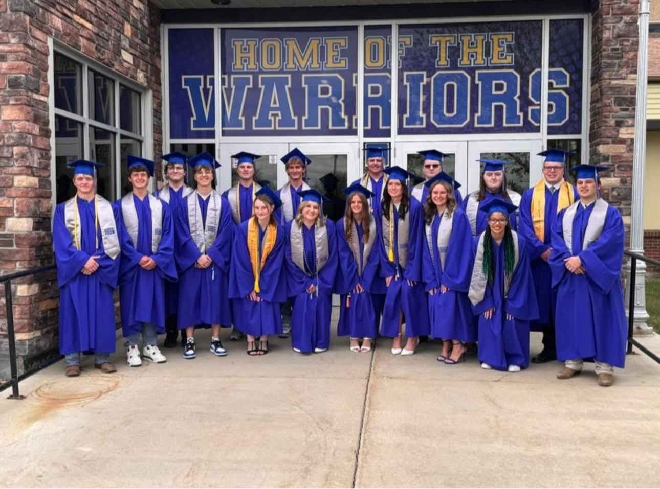 Group of students in blue gowns and caps standing in front of a brick building.