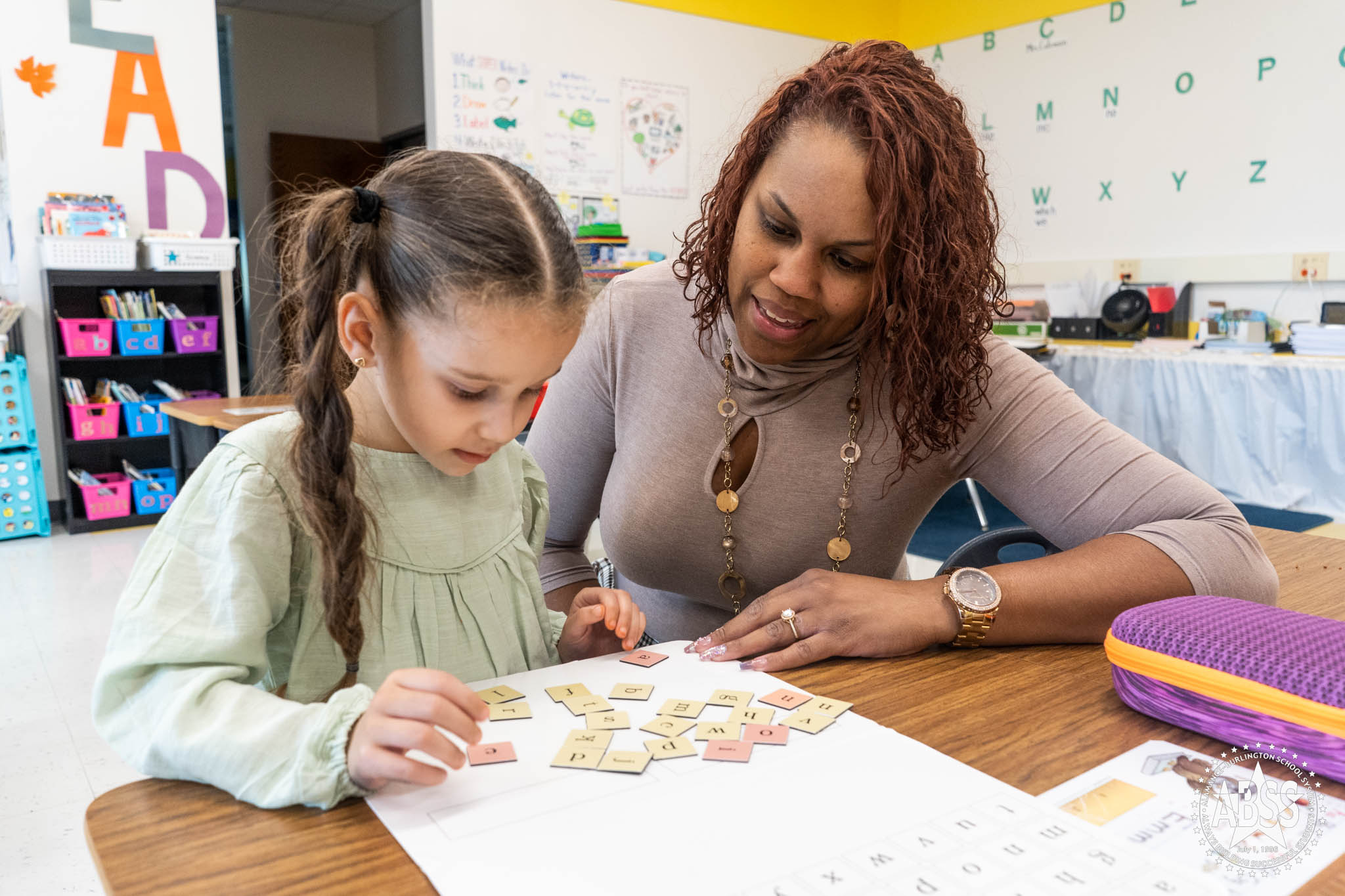 An elementary teacher sits beside a student in a green sweater as she assembles word tiles on a whiteboard