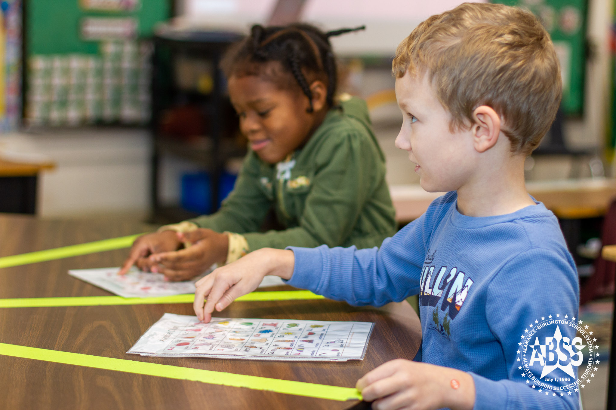 Two kindergarten students are working at a brown table. The boy in the foreground in a blue shirt is pointing at a word on the board and looking up at the teacher. The girl in the background is looking down and making an inquisitive face.