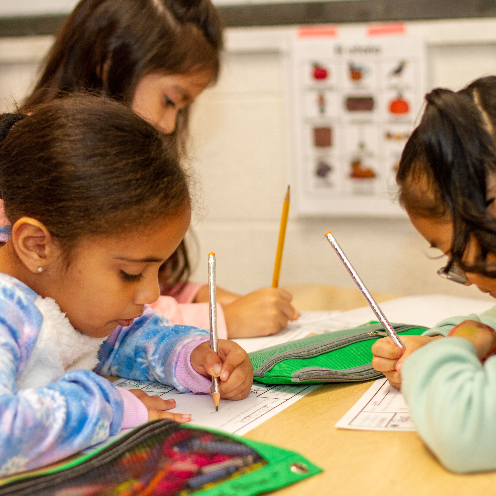 Three Kindergarten girls looking down at their paper assignment while they hold a pencil at a small group table.  There is a green pencil pouch on the table with them.