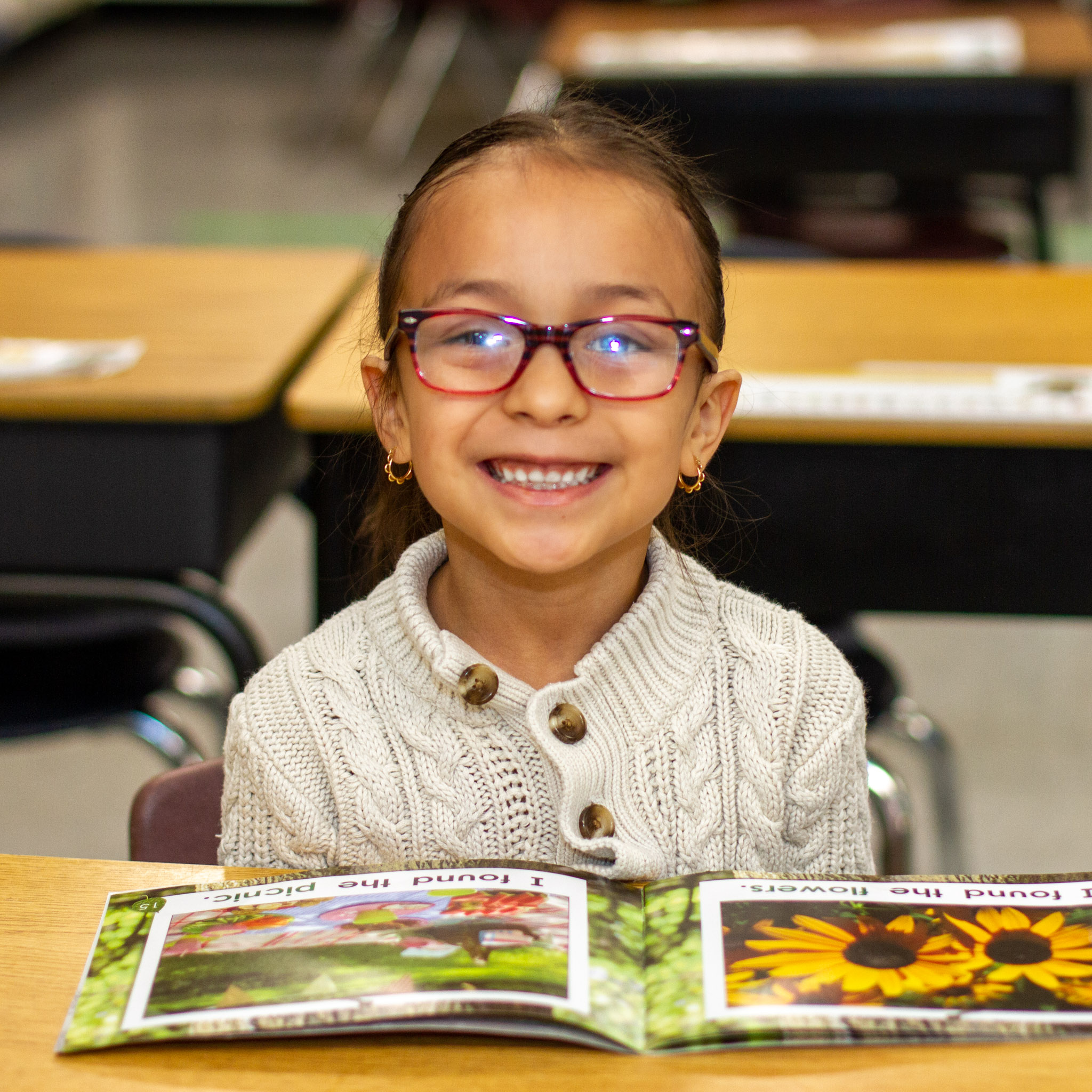 A kindergarten girl in a white button up top and glasses smiles at the camera with an open picture book on her desk