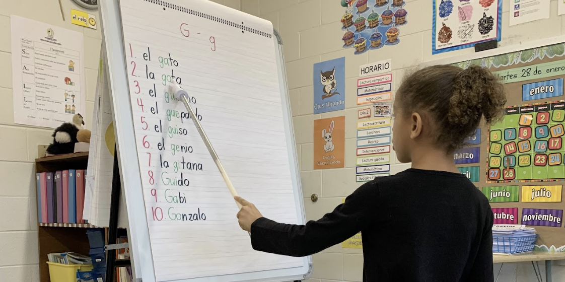 A young girl points at "la gata" on lined chart paper using a pointer