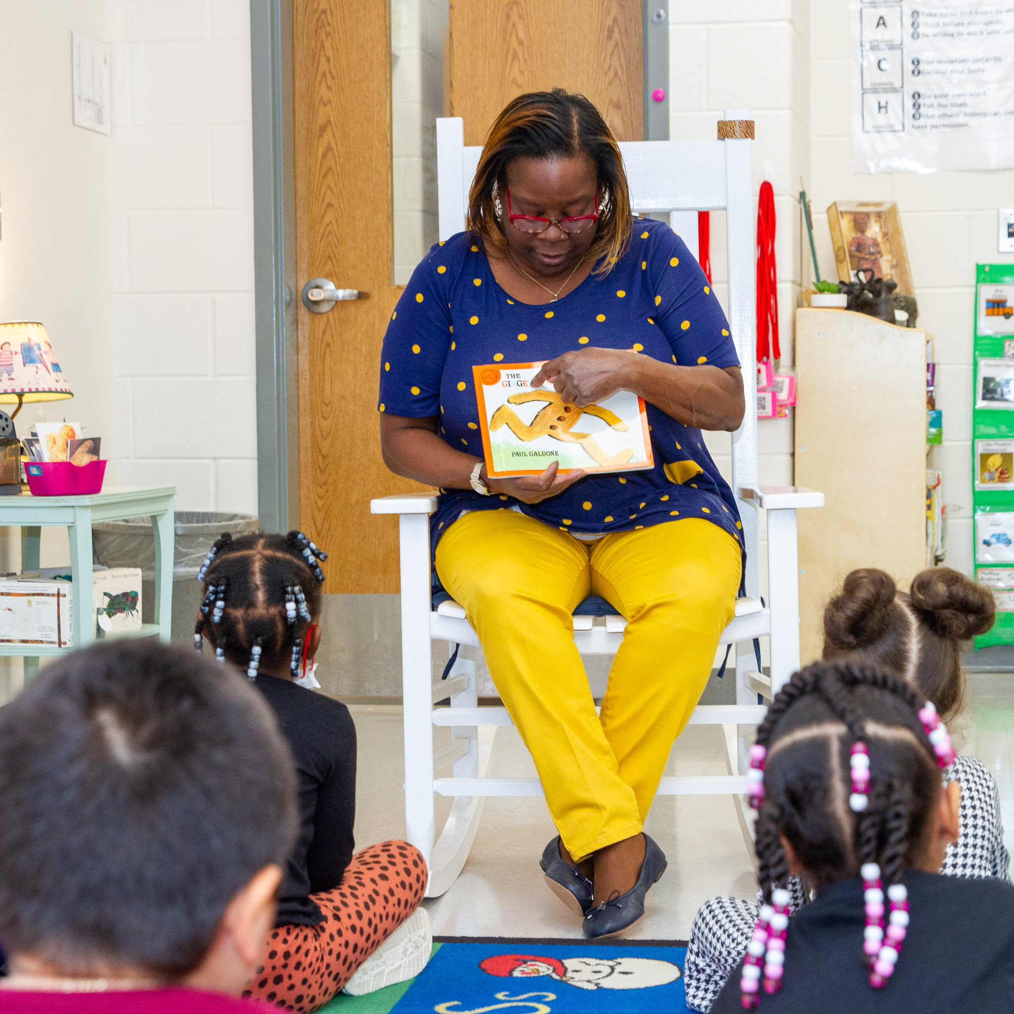Teacher wearing yellow pants and blue shirt, sitting in a white rocking chair,  points to the cover a children's book with students looking on the carpet