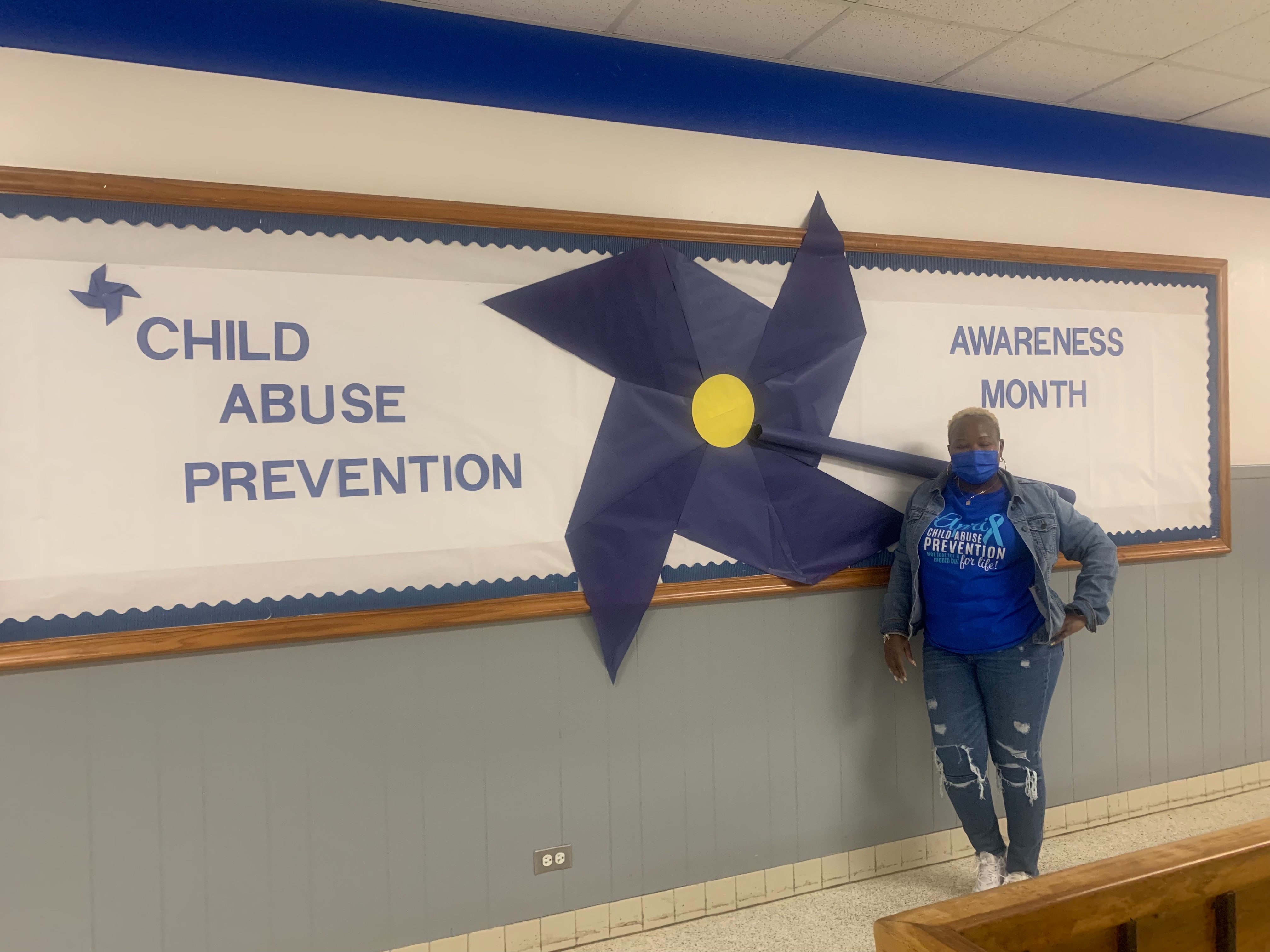 School Social Worker standing in front of bulletin board that says Child Above Prevention Awareness Month and a large cutout blue flower