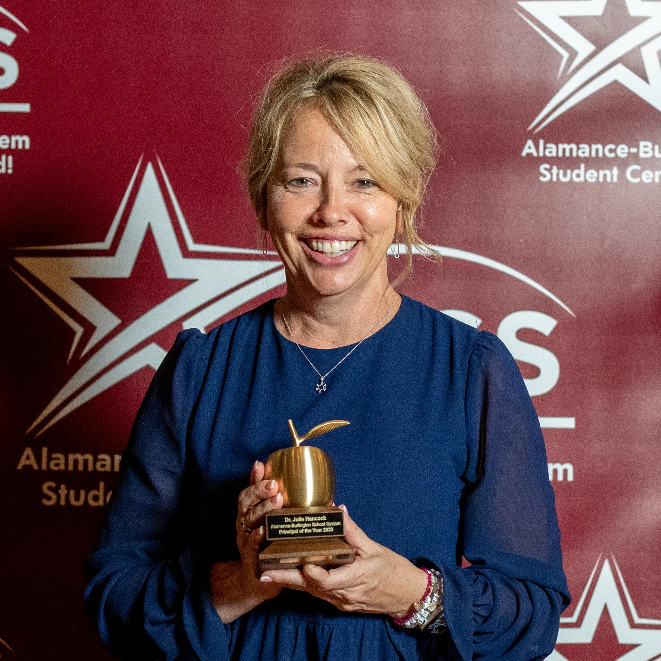 Dr. Julie Hancock, ABSS Principal of the Year holding Golden Apple Trophy in front of a burgundy ABSS background