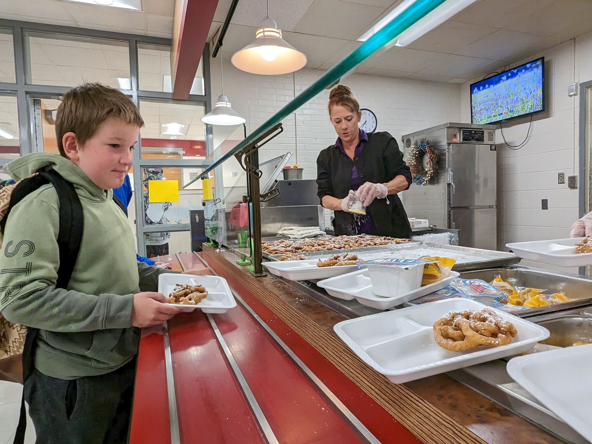 A young student slides his breakfast tray down the cafeteria counter as the nutrition specialist greets him