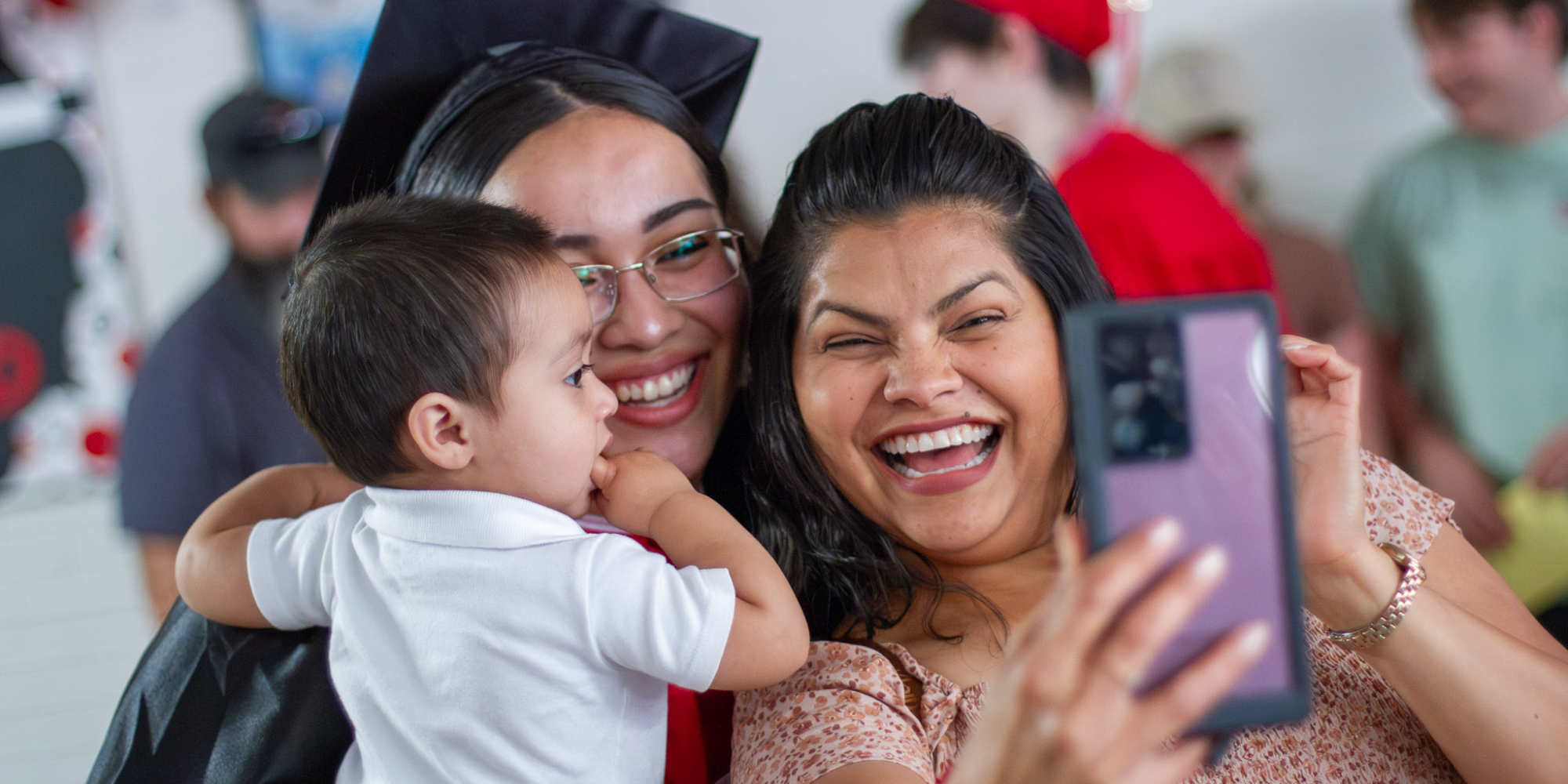A parent takes a selfie with her graduating senior child in cap and gown while she holds a toddler