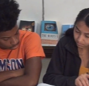 Two students looking on at a book between them on a desk