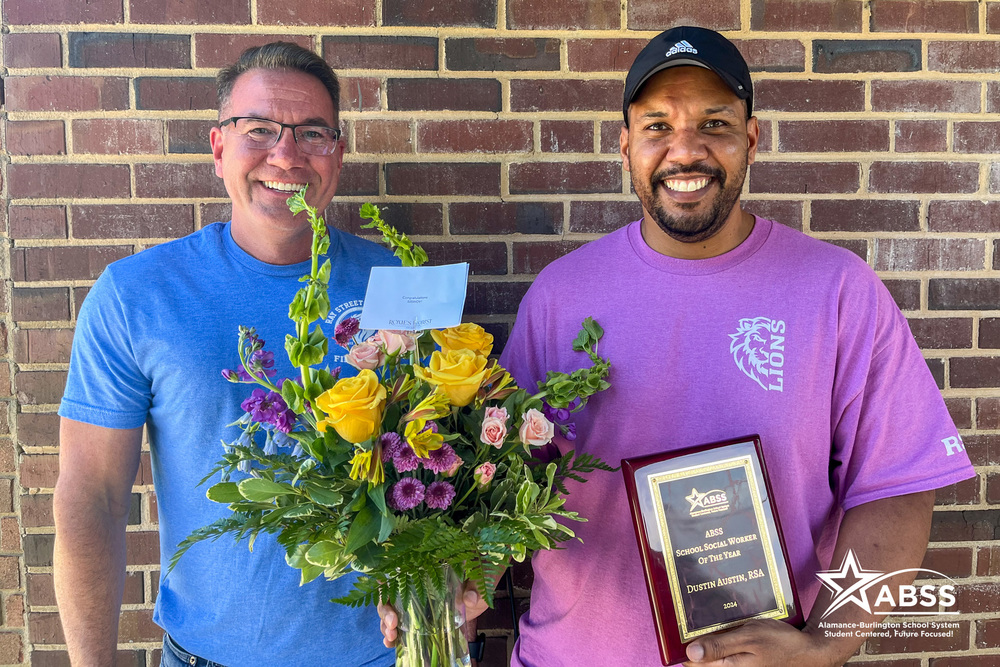 Dustin Austin, School Social Worker of the Year, standing outside holding flowers and plaque.  Dr. Bayless, Principal standing beside Dustin.