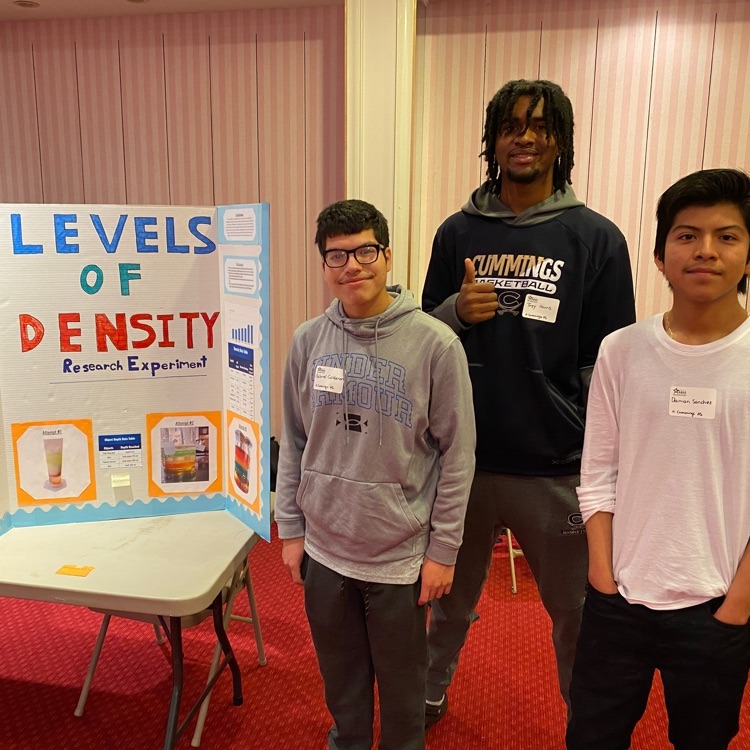 Three teenage boys stand in front of a science fair project board titled "LEVELS OF DENSITY Research Experiment". The board shows images of liquids with different densities layered in glasses. The boys appear to be of diverse backgrounds and are smiling at the camera.