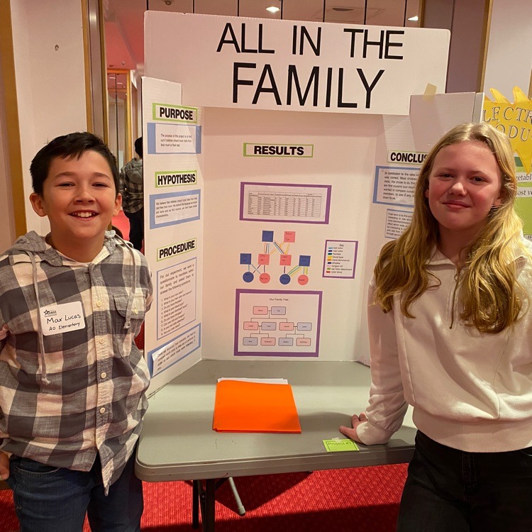 Two students, a boy and a girl, stand on either side of a science fair project board titled "ALL IN THE FAMILY". The board shows various charts and diagrams related to family genetics. Both students are smiling at the camera.