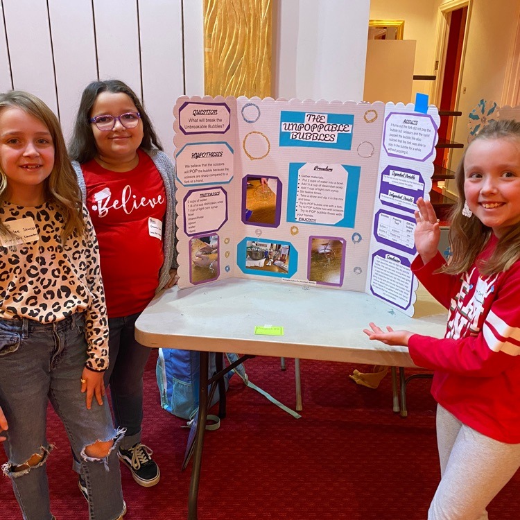 Three young girls pose in front of a science fair project board titled "The Unpoppable Bubbles". They're smiling and appear excited about their project. The board has sections for the question, hypothesis, procedure, and results of their bubble experiment.