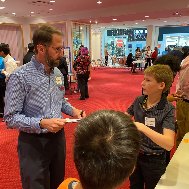 An adult man with glasses and a beard, wearing a blue striped shirt, is engaged in conversation with a young boy in a dark polo shirt. They appear to be at a science fair or similar event, with other people and displays visible in the background. The setting has red carpeting and decorative lighting. The man is holding papers, suggesting he may be evaluating or discussing the boy's project.