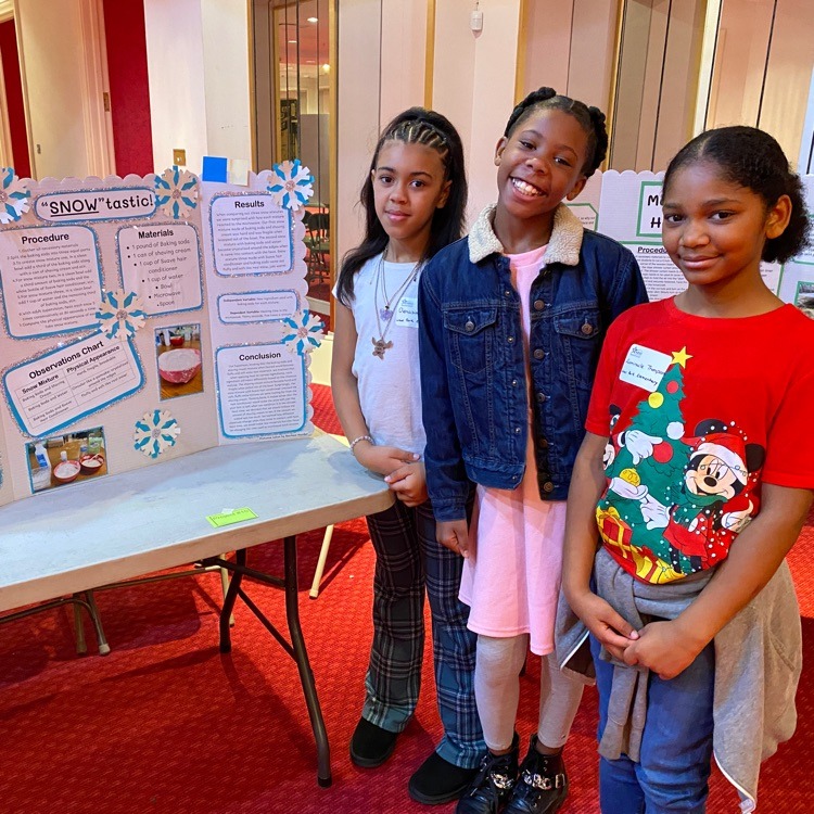 Three girls pose next to a science fair project board titled "'SNOW'tastic!". The board has a winter theme with snowflake decorations. It includes sections on procedure, materials, results, and conclusions for what appears to be a snow-related experiment.