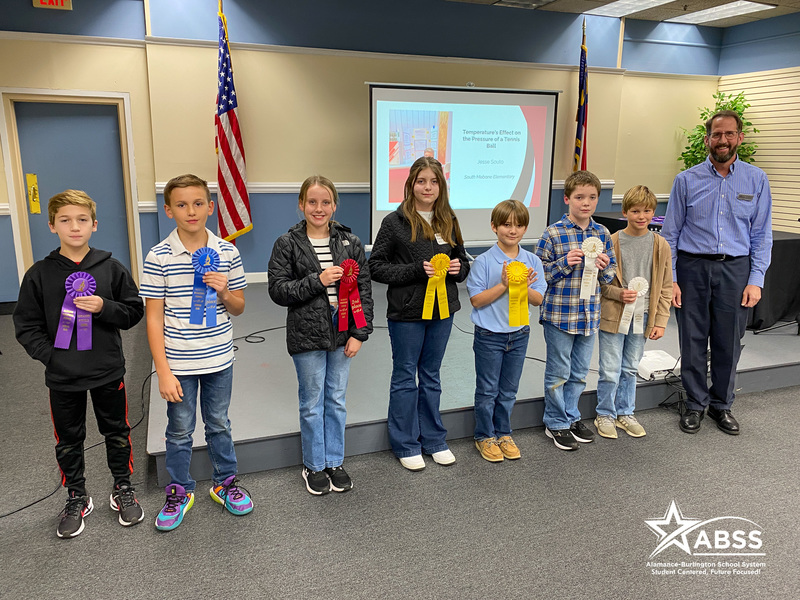 Seven students and an adult man stand in a line, each holding a colorful ribbon award. Behind them is a projection screen and two American flags. The students appear to be of various ages and genders. The image has an "ABSS" logo in the bottom right corner.