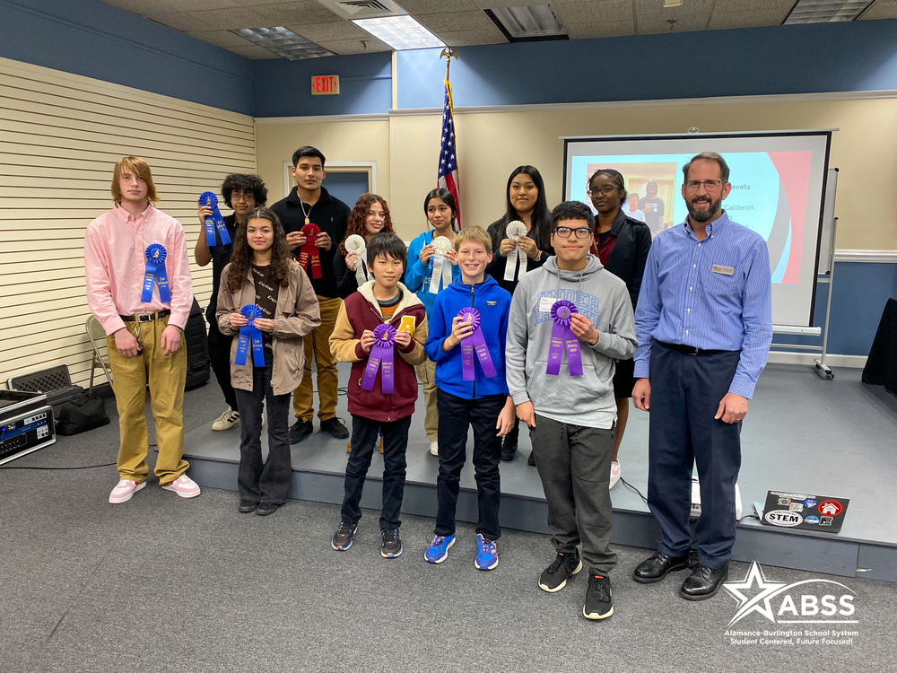 A group of about 12 students and an adult man pose on a small stage. They are holding various colored ribbon awards. Behind them is a projection screen and an American flag. The students are diverse in age, gender, and ethnicity. The image has an "ABSS" logo in the bottom right corner.