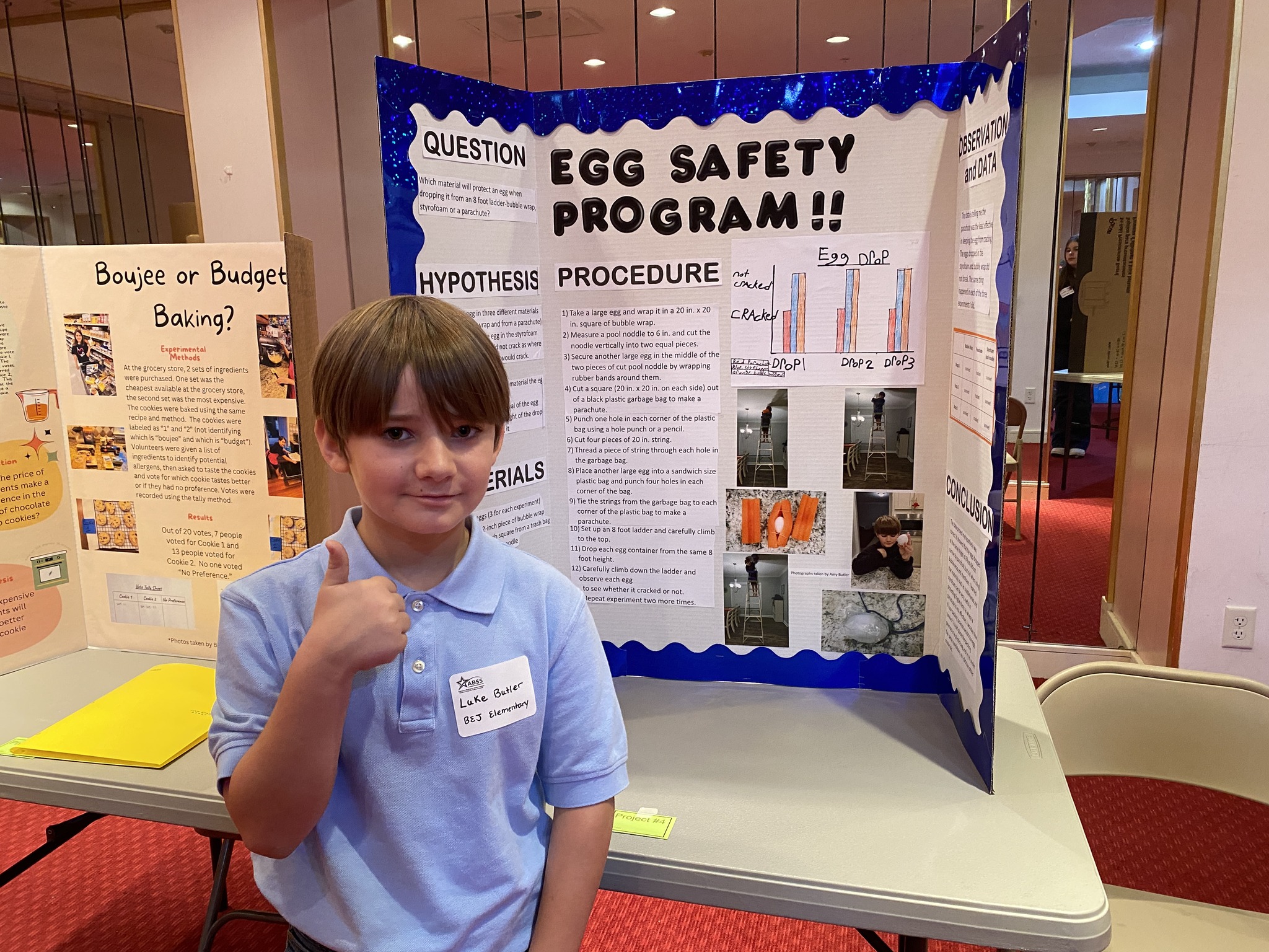 A young boy with short hair stands in front of a science fair project board titled "Egg Safety Program!!". He's wearing a light blue polo shirt and smiling while giving a thumbs up. The project board details a hypothesis, procedure, and results related to egg safety experiments.