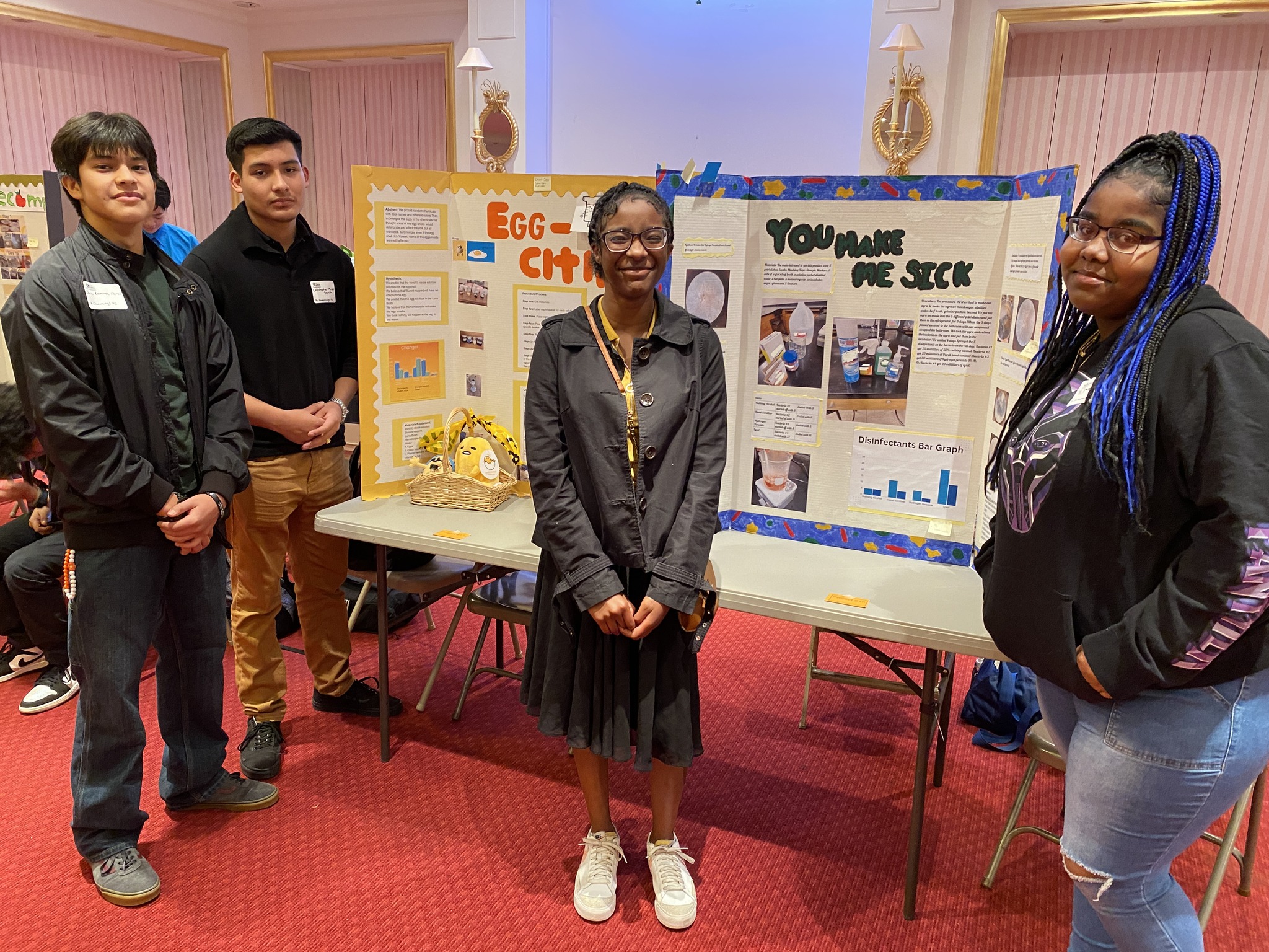 Four students stand in front of science fair project boards. Two project boards are visible, titled "Egg City" and "You Make Me Sick". The students appear to be of diverse backgrounds. The setting is an indoor room with pink curtains and decorative wall sconces.