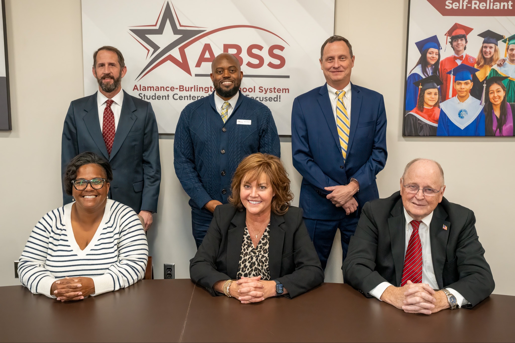 Group photo of six ABSS (Alamance-Burlington School System) board members positioned in front of an ABSS logo wall. Three members are seated in the front row at a dark wood conference table, while three stand behind them. The ABSS logo features a star design with "Student Centered, Future Focused!" tagline. The members are professionally dressed, with the men wearing suits and ties, one woman wearing a striped sweater, and another in business attire with a leopard print blouse. A graduation photo display is visible on the right side of the wall. The overall setting appears to be a professional conference or meeting room.