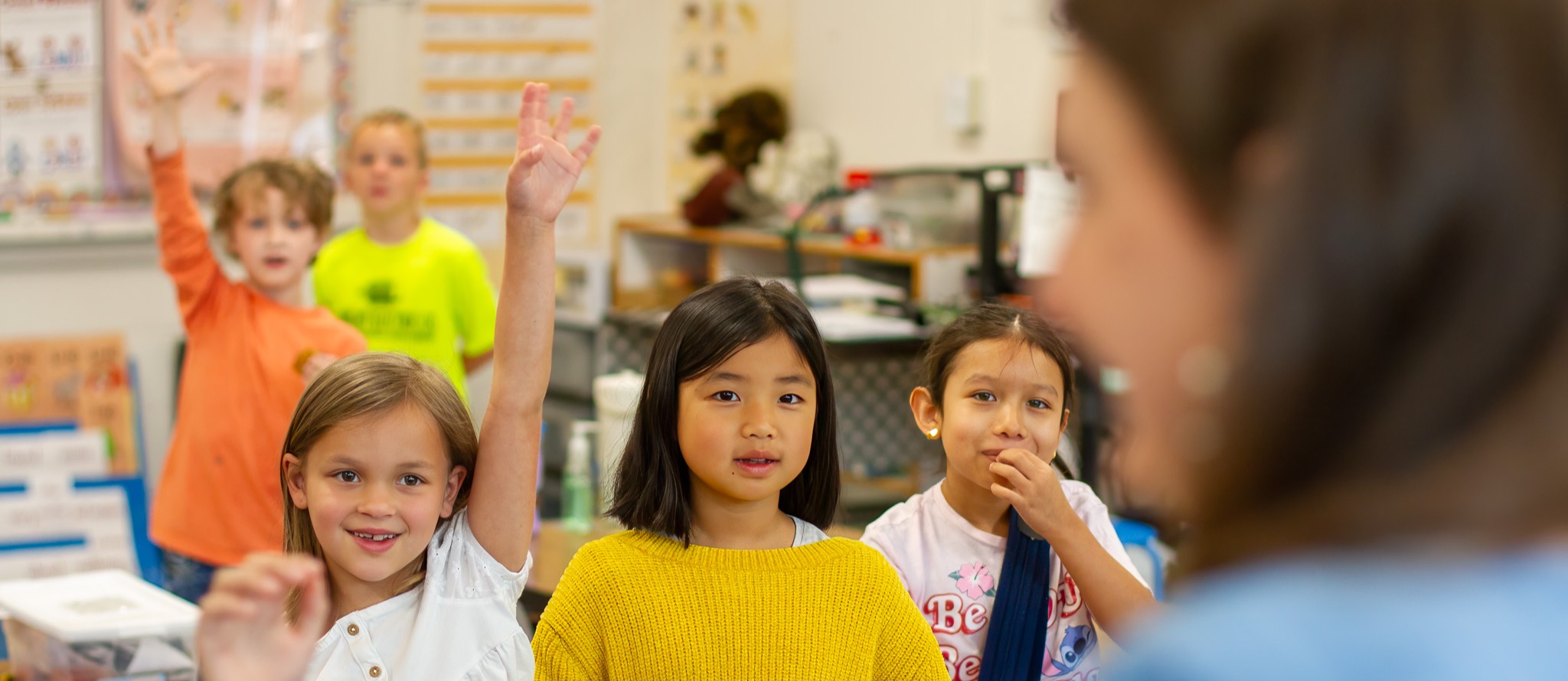 A diverse group of elementary school children in a classroom. Several students are raising their hands, with one girl in a white shirt prominently raising her hand in the foreground. A girl in a yellow sweater holds a green object in the center. The classroom background shows educational posters and materials.