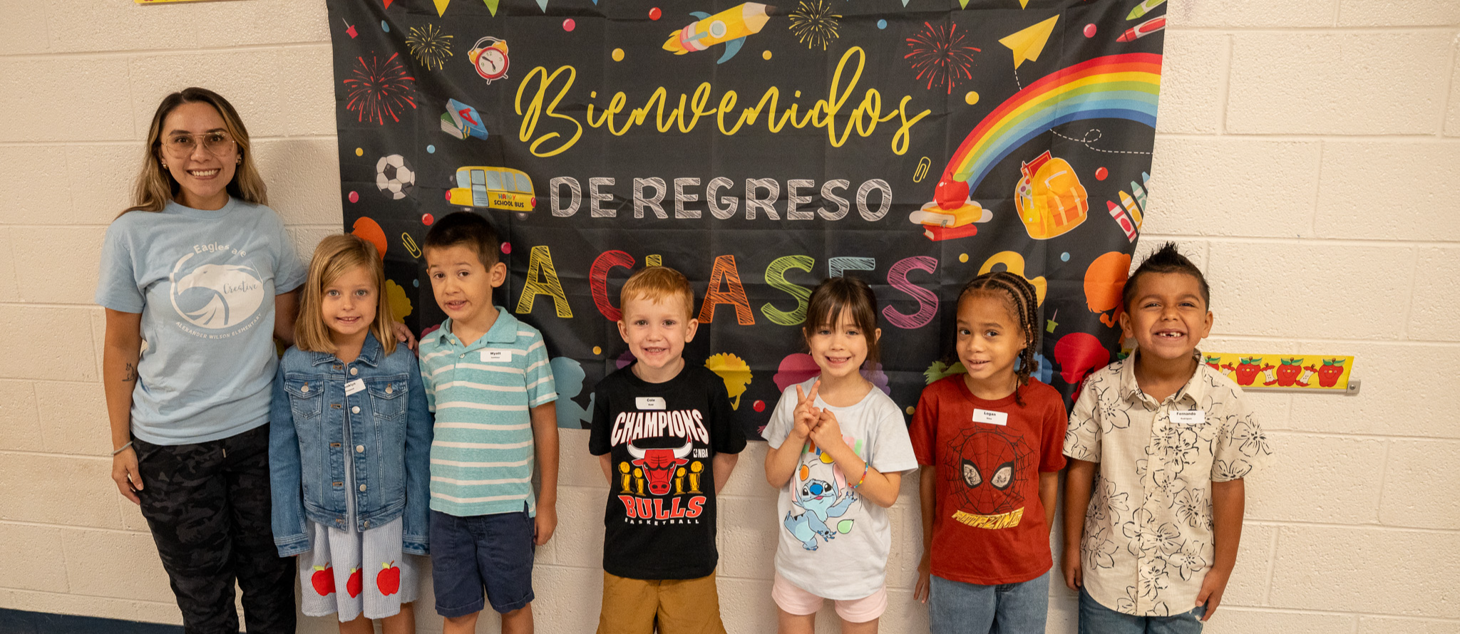A class of multilingual students standing beside their teacher in front of a vibrant multicultural bulletin board