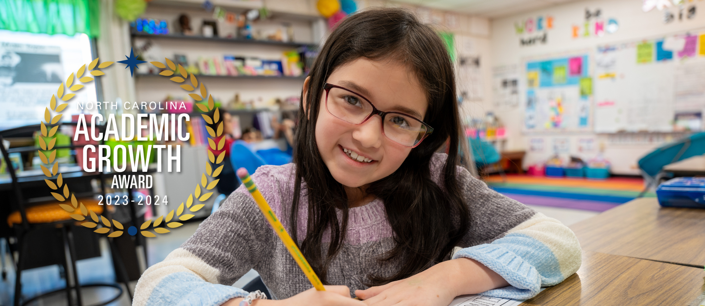 Photo of a student smiling while holding a pencil working at her desk with a medal award showing school met academic growth to the left side