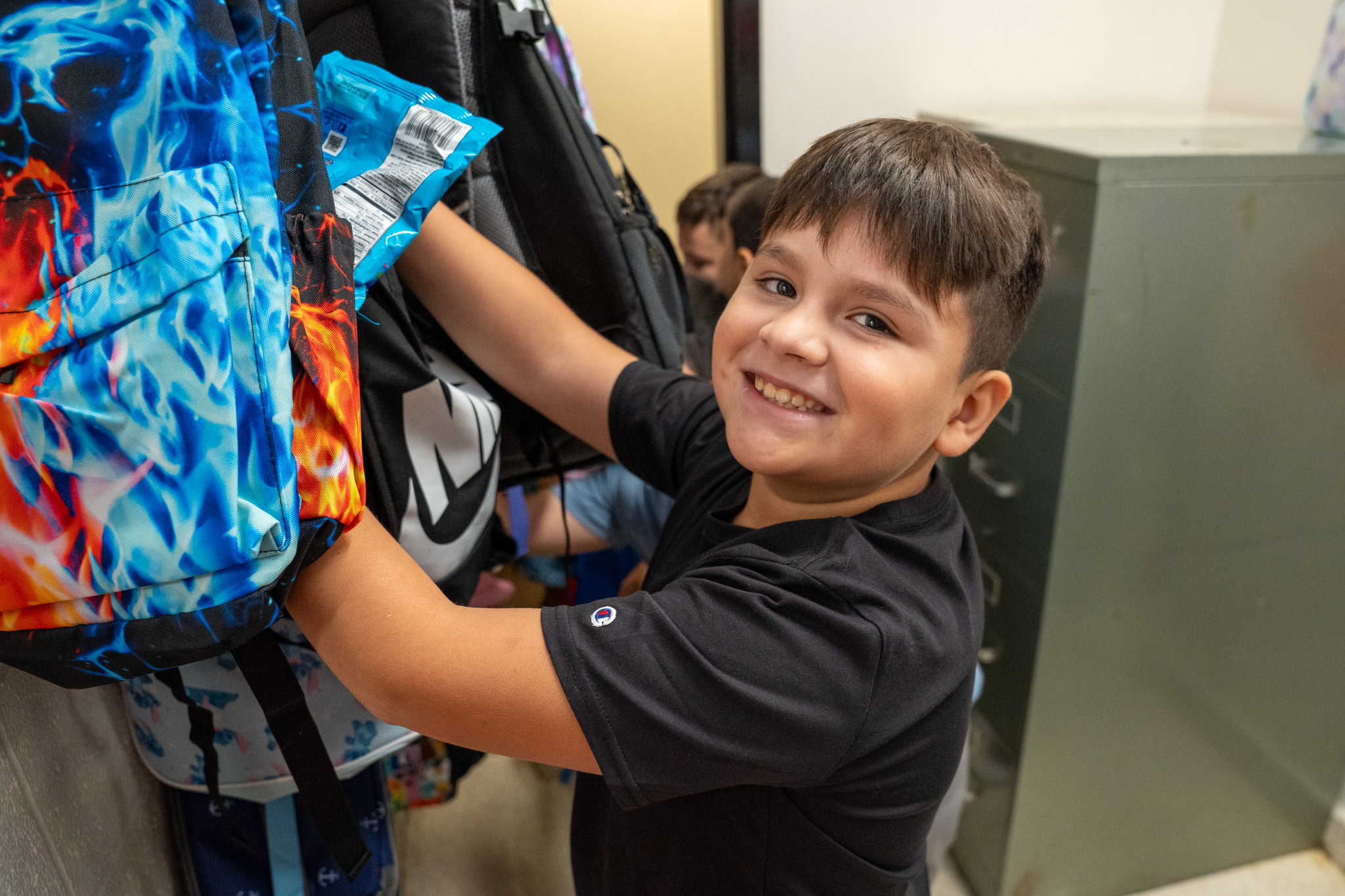 A young boy hanging up his backpack on a hook and looking towards the camera smiling