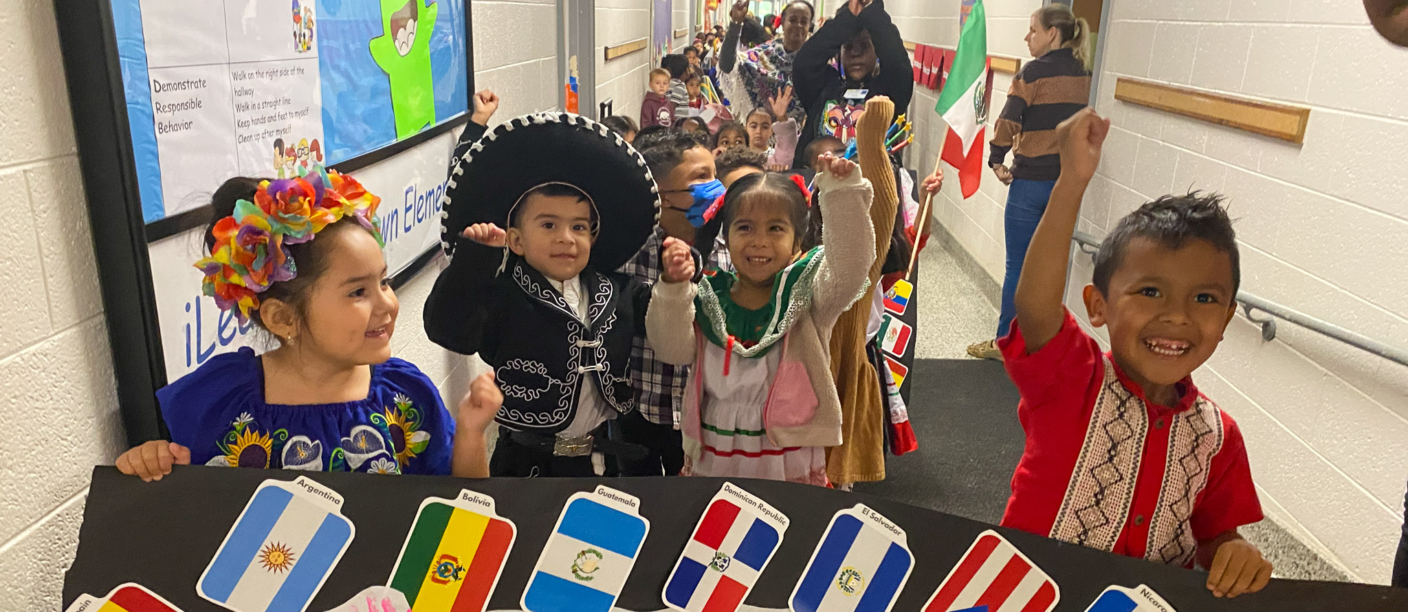 Pre-K students hold up a banner that celebrates Hispanic cultures while a line of more students stands behind them and cheers