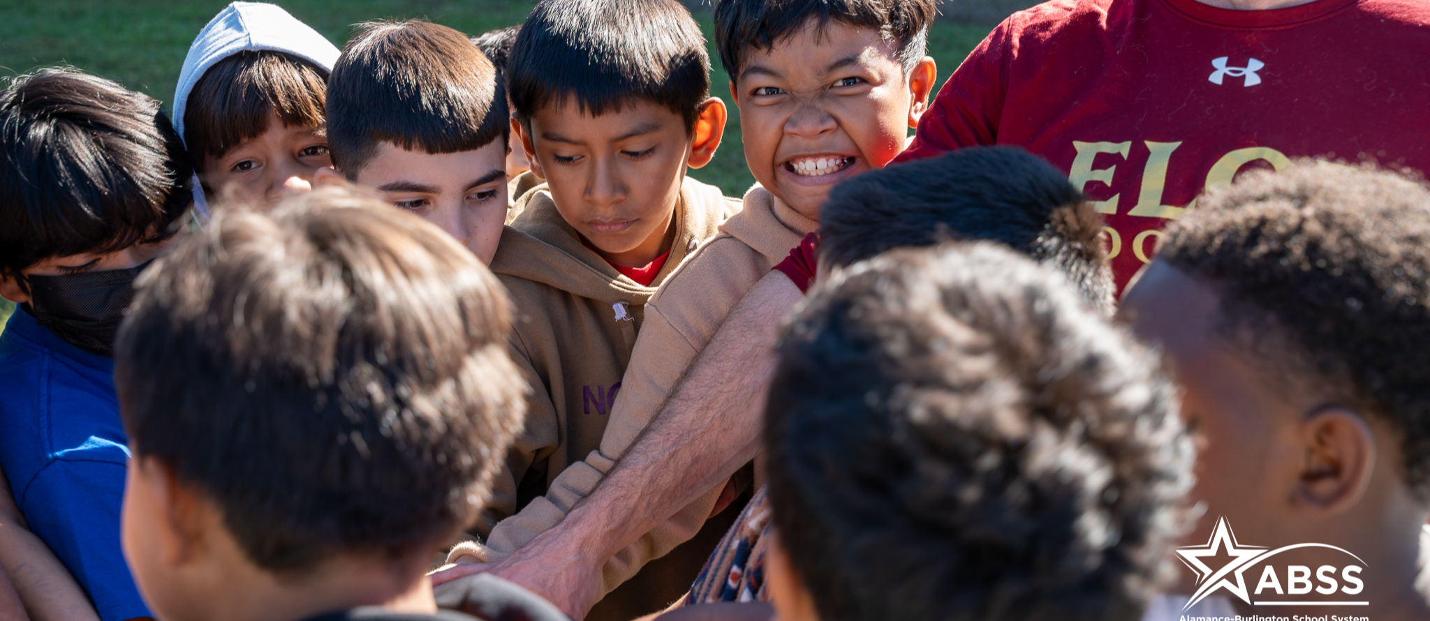 Elementary boys huddle together in a high five formation with an adult wearing an Elon Football shirt joining in from the side