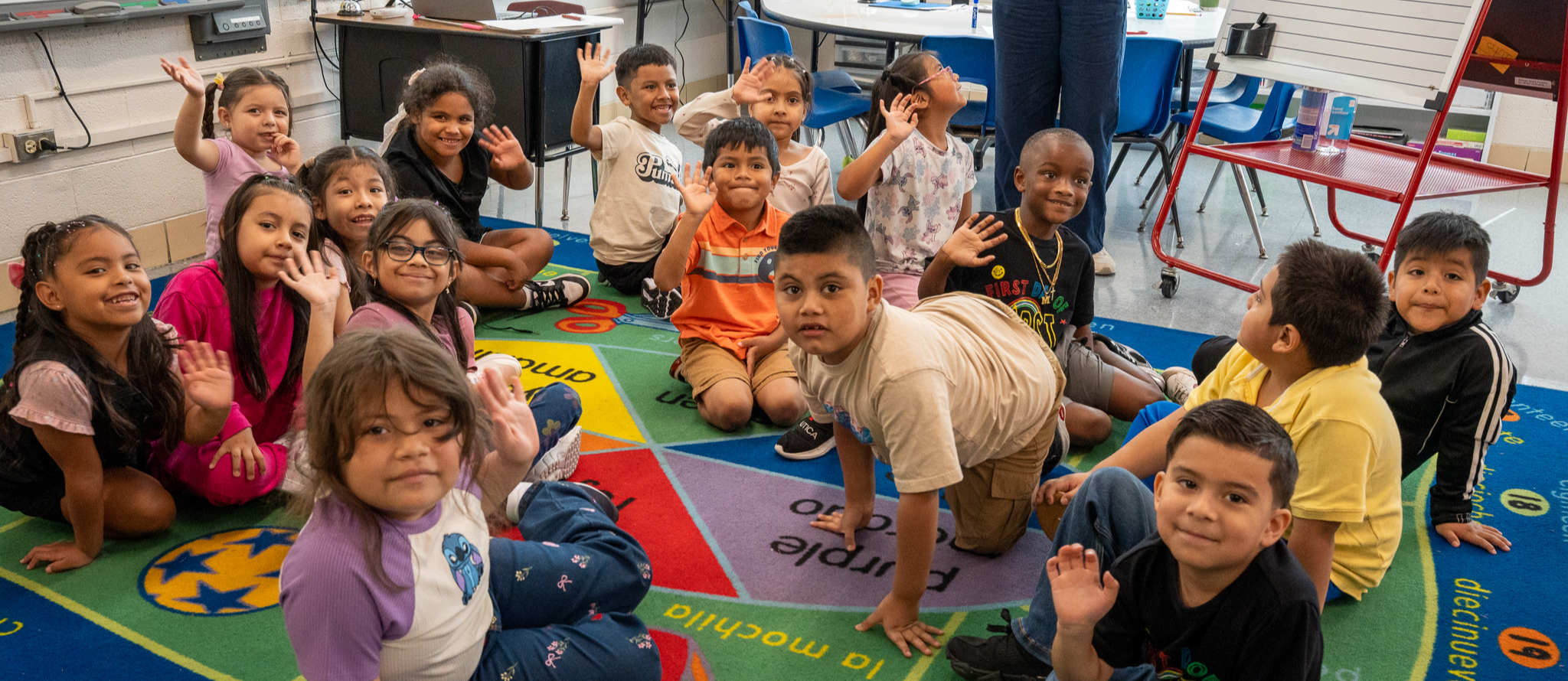 A class pauses on a classroom carpet to look up at the camera and wave
