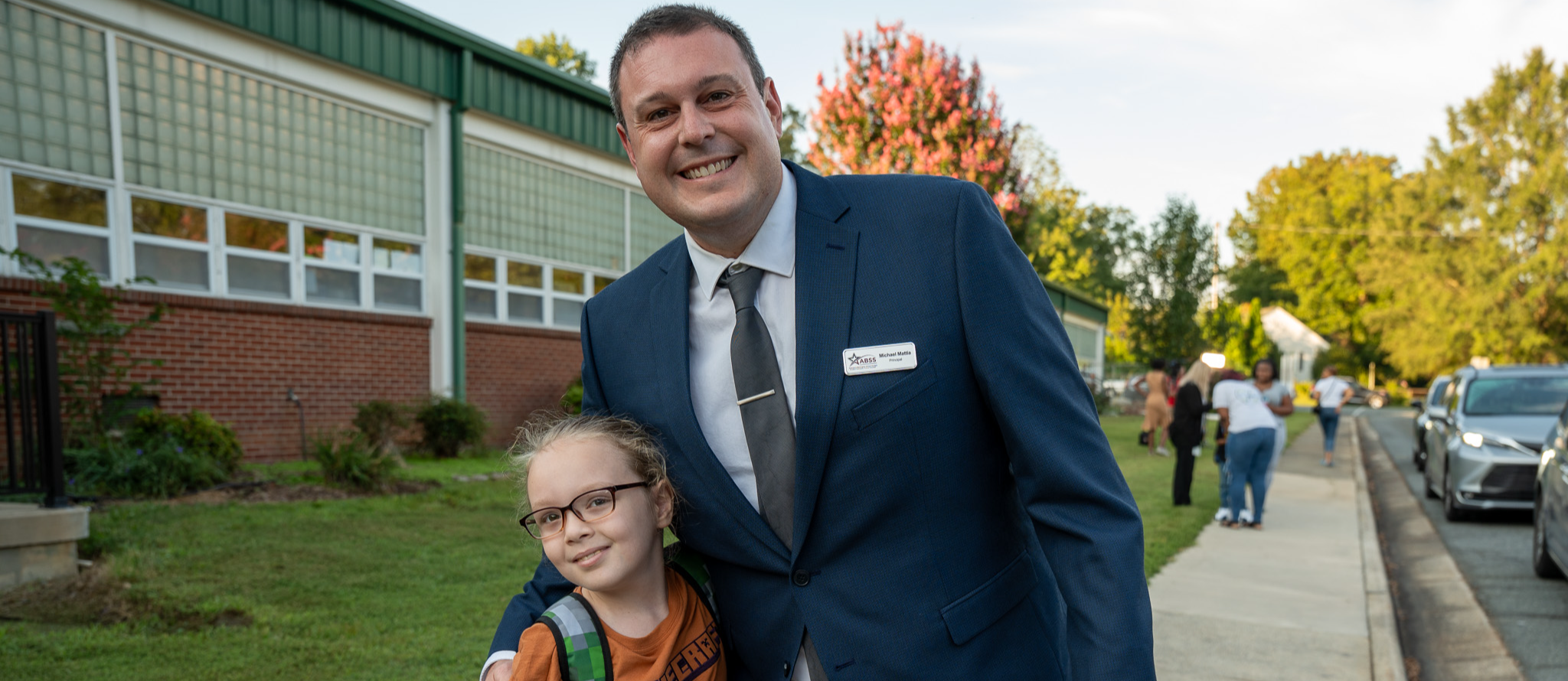 Principal wearing a suit gives a side hug to a young girl who just got out of the car in the morning line