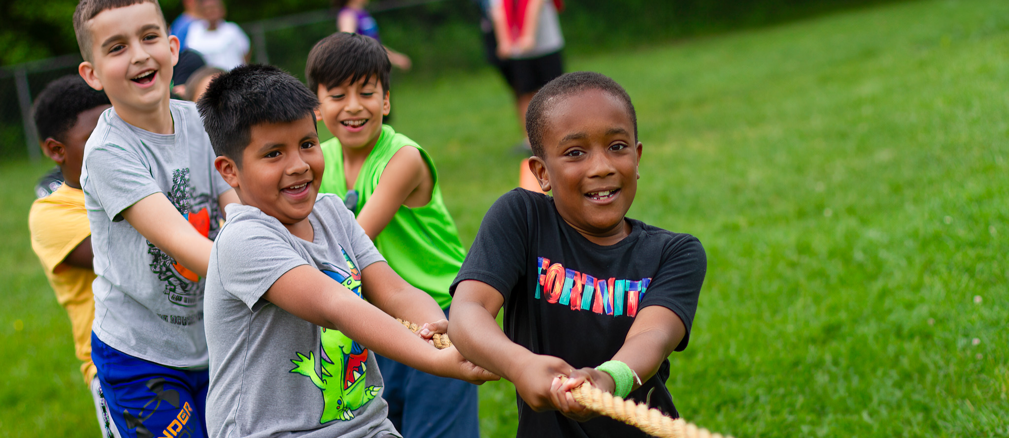 A group of diverse children play tug-of-war on a grassy field, smiling and laughing. Four boys in the foreground pull on a rope together, wearing colorful t-shirts and shorts. Other children and a grassy area with trees are visible in the background. The image captures a moment of teamwork and joy during an outdoor activity.