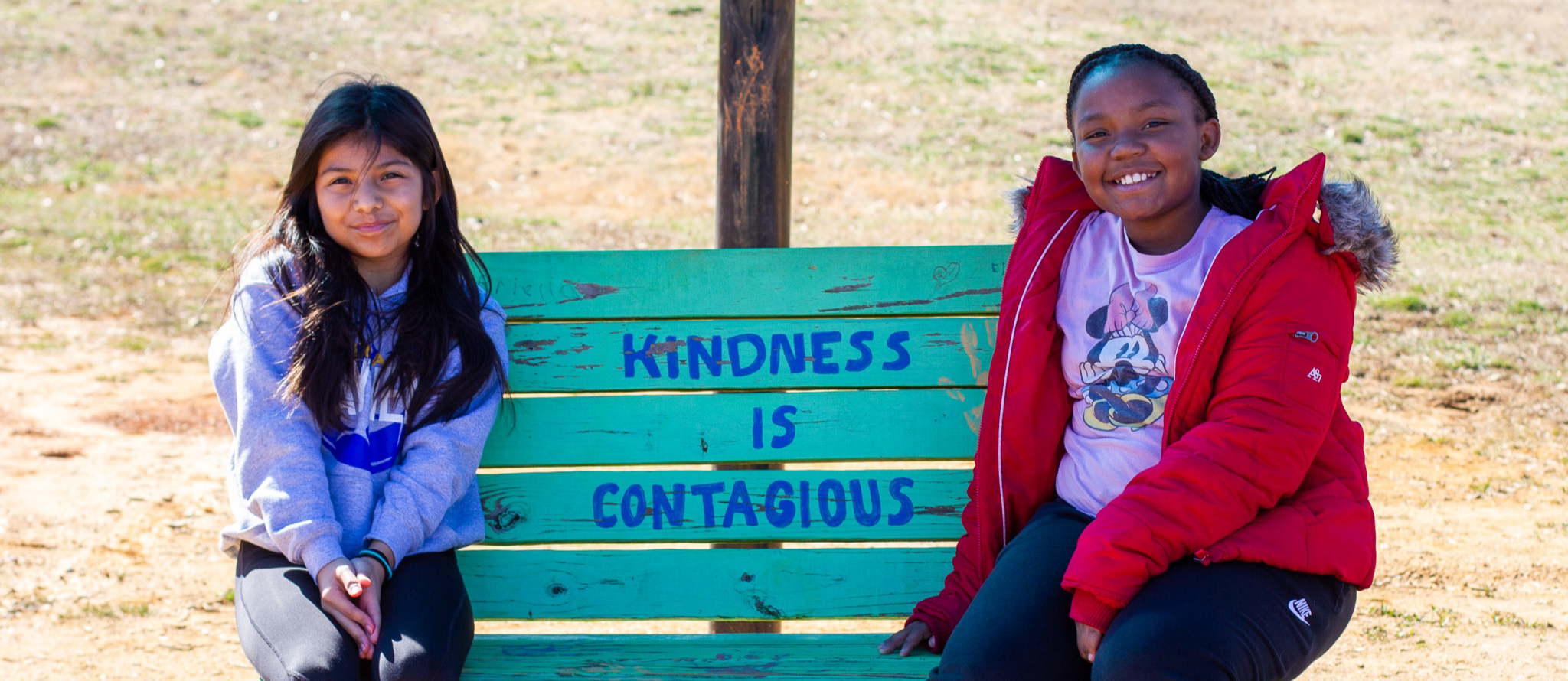 Photograph of two fifth grade girls at Highland Elementary sitting on a bench on the playground with the words "Kindness is Contagious" painted on the bench showing
