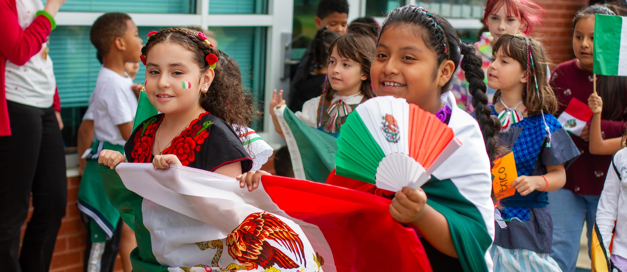 Children celebrating Mexican culture at Highland Elementary School. Two girls in the foreground hold a large Mexican flag and a tricolor fan. They wear traditional Mexican attire and have colorful decorations in their hair. Behind them, other children hold flags and wear festive clothing. The scene takes place outside the school building on a sunny day.