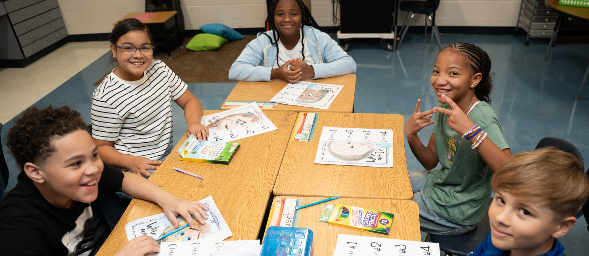 Five students seated at a cluster of desks look up from a group activity