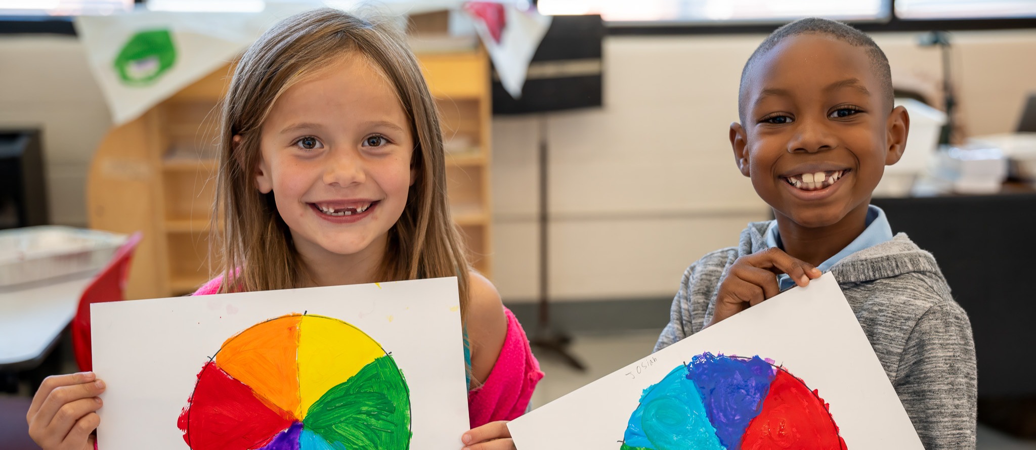 Two elementary school children, a girl with long blonde hair and a boy with short dark hair, smiling and holding up colorful color wheel paintings they created. The classroom setting is visible behind them.