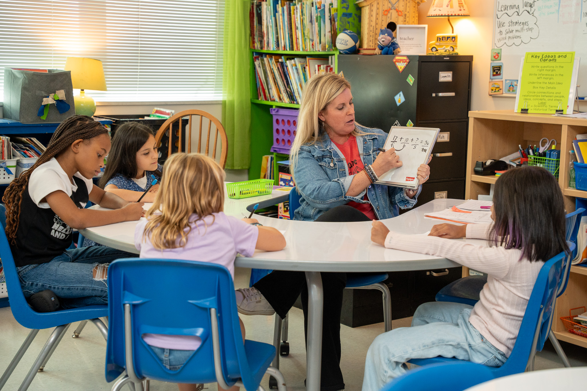 A teacher in a denim jacket leads a group of four students in a math lesson at a small group table