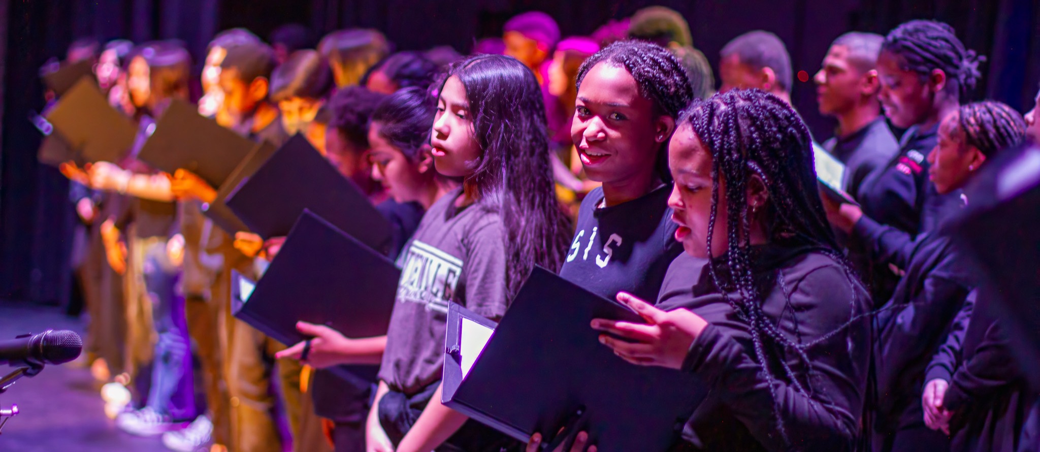 A diverse group of students stands on stage in a choir formation, holding black folders. They are illuminated by purple and orange stage lighting. The students appear to be singing or performing, with some looking at their folders and others facing forward. The image captures a mix of ethnicities and ages among the performers. In the foreground, several students are in focus, including young women with long hair and braids. The ABSS (Alamance-Burlington School System) logo is visible in the bottom right corner of the image.