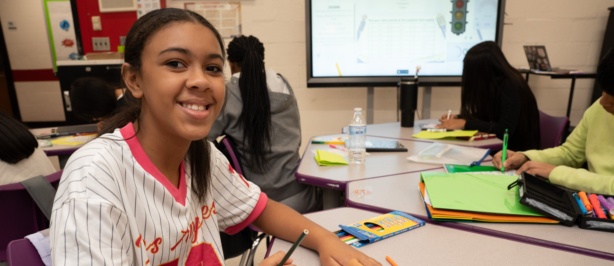 A teenage girl in a white baseball jersey looks up from her work at her desk in class