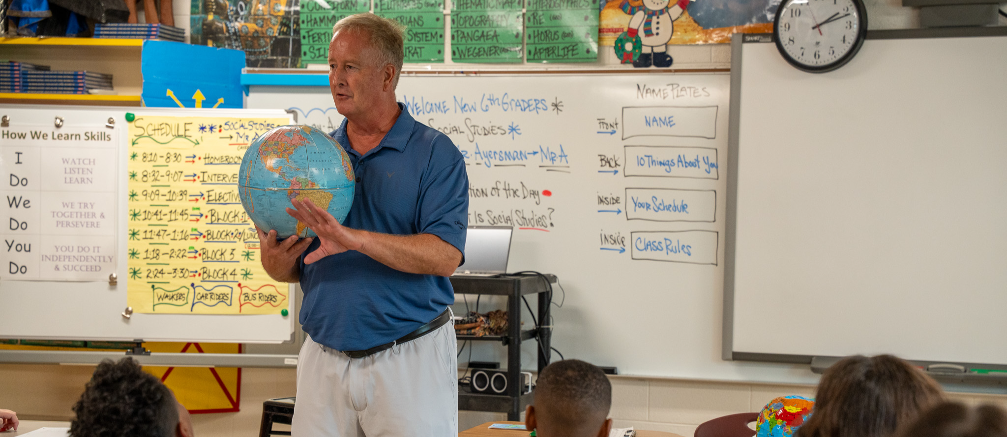 A male teacher wearing a blue shirt and khaki pants holds a globe in front of his middle school social studies classroom